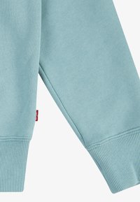 Manga de um sweatshirt azul claro, com punho canelado e textura suave. Uma pequena etiqueta vermelha é visível perto da costura.
