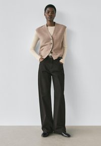 Beżowa dzianinowa kamizelka z dekoltem w serek, zapinana na guziki; pod spodem długi beżowy top. Brązowe spodnie o szerokich nogawkach i czarne, spiczaste buty. Gładka faktura.