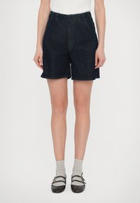 Shorts en denim foncé à taille haute, avec des coutures vertes, des plis et des ourlets roulés, associés à des chaussettes grises et des chaussures marron.