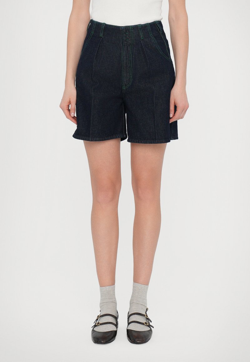 Shorts en denim foncé à taille haute, avec des coutures vertes, des plis et des ourlets roulés, associés à des chaussettes grises et des chaussures marron.