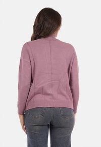 Donna che indossa un maglione a dolcevita color malva e jeans blu scuro, in piedi con la schiena rivolta verso la fotocamera su uno sfondo neutro.