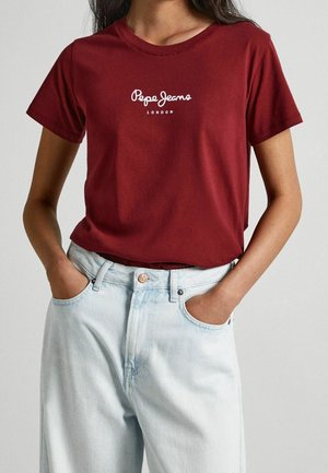 Person trägt ein bordeauxrotes Pepe Jeans London T-Shirt, das in eine hellblaue, hoch taillierte Jeans gesteckt ist, mit den Händen in den Taschen.