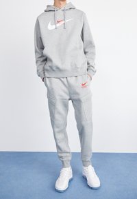 Šedá mikina s bílým logem Nike a červenými akcenty, doplněná o odpovídající šedé tepláky s cargo kapsami a bílými teniskami.