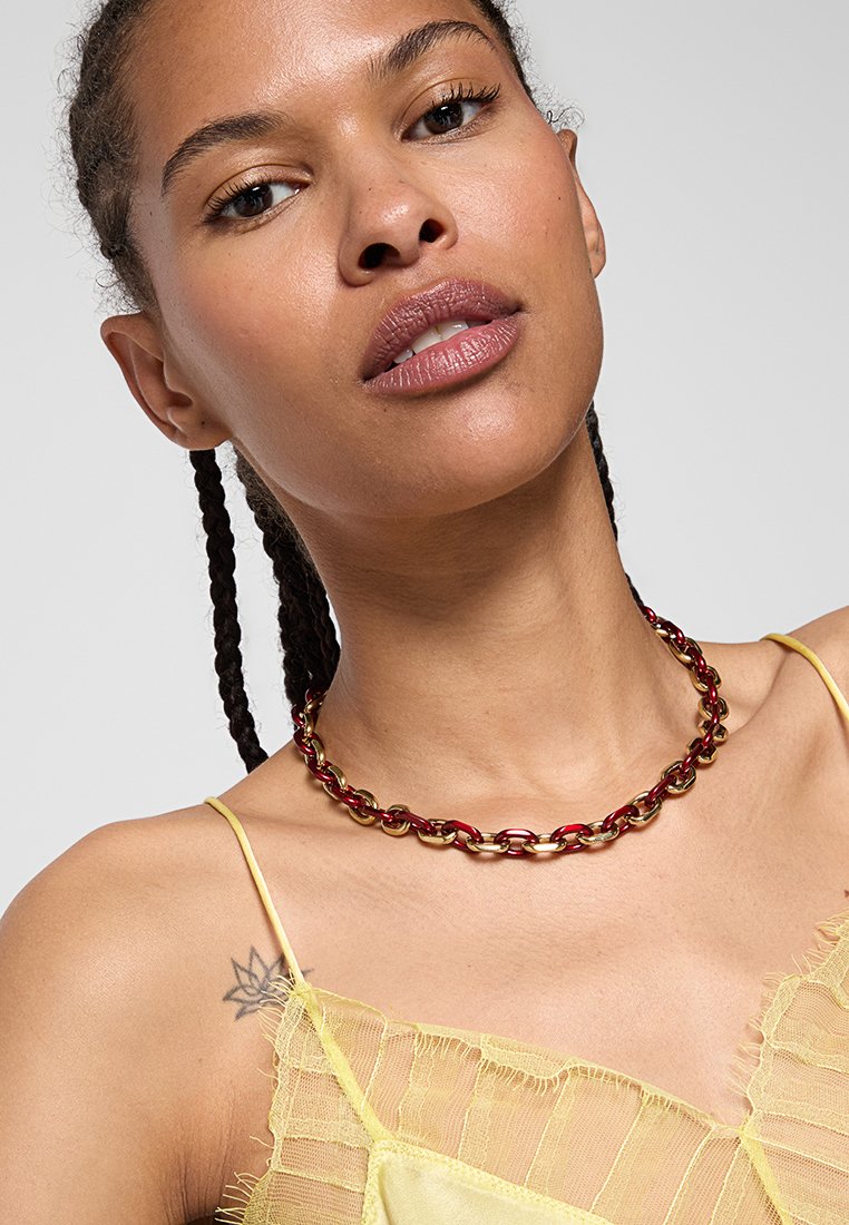 Czerwono-złoty łańcuszek, składający się z połączonych owalnych ogniw. Noszony na obnażonej szyi, uzupełniony przez przezroczystą żółtą bluzkę.
