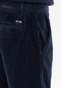 Granatowe sztruksowe spodnie w pionowe paski, z bocznymi kieszeniami i metalowym logo z tyłu. Miękka faktura, dopasowany krój.