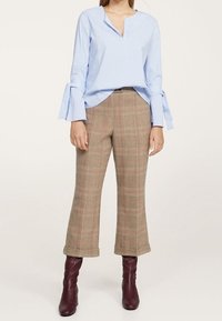 Chemise bleu clair à manches longues avec un col en V et des poignets évasés, associée à un pantalon large à carreaux marron et des bottines bordeaux.