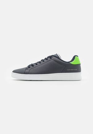 Czarny niski sneaker z białą podeszwą, czarnymi sznurowadłami i neonowo zieloną taśmą na pięcie, z logo Tommy Hilfiger po boku.