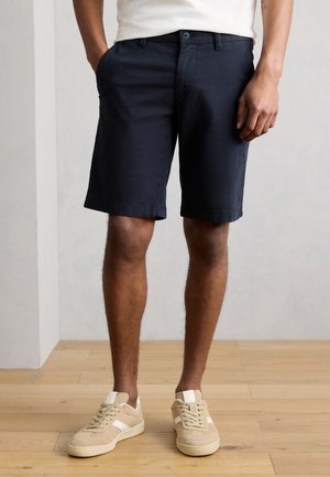 Mand iført marineblå shorts, hvid skjorte og beige sneakers, stående på trægulv mod en ensfarvet lys væg.