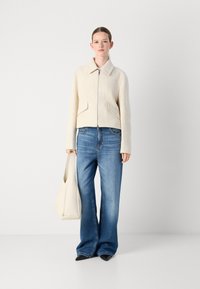 Beige textuurgekapte jas met een kraag en een ritssluiting aan de voorkant, gecombineerd met een wijde blauwe jeans en een crèmekleurige handtas.