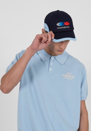 Mann richtet marineblaue und hellblaue Kappe mit "PROHIBITED"-Logo, trägt hellblaues Polohemd mit passendem Schriftzug "PROHIBITED Racing Club".