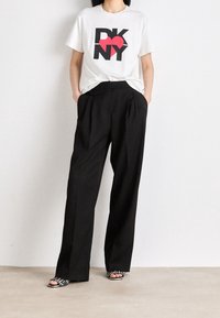 Biała koszulka z czarnym i czerwonym logo "DKNY", zestawiona z czarnymi spodniami o wysokim stanie i szerokich nogawkach oraz wzorzystymi sandałami na otwartym nosku.
