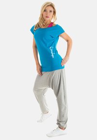 Blue oversized top with side logo text, paired with light gray harem pants. Model wears white sneakers, highlighting a relaxed, casual look.