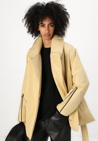 Beige oversized jakke med høj krave, lynlåslåse ærmer, glat stof og løs pasform, parret med en sort top og læderbukser.