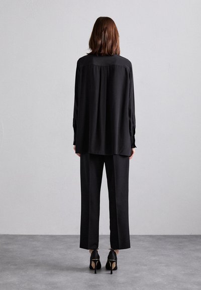 Black blouse and loose-fitting black trousers, both in a smooth fabric. The blouse has long sleeves and a collar. High-heeled black shoes.