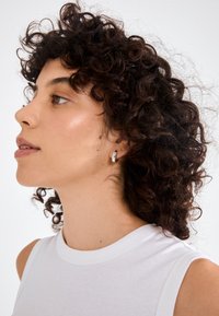 Sidabrinė apyrankė ant modelio su garbanotais plaukais, vilkinčioje be rankovių baltoje bluzonėje. Neutralus fonas išryškina papuošalo dizainą.