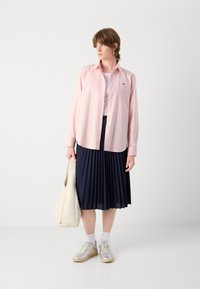 Camicia rosa chiaro a maniche lunghe, gonna plissettata blu navy, sneakers bianche con dettagli argentati e borsa a tracolla beige. Tessuto morbido e vestibilità comoda.