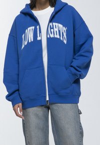 Felpa con zip blu, con il testo bianco "LOW LIGHTS", tasche frontali e cappuccio con cordino. Polsini e orlo a coste, realizzata in tessuto morbido.