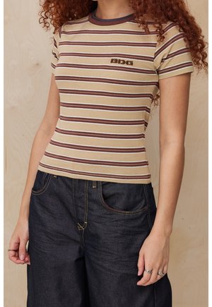 Kobieta ubrana w beżowo-brązową koszulkę w paski z krótkim rękawem z logo "BDG" oraz ciemne spodnie dżinsowe, stojąca na jednolitym tle.