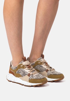 Gambe con indossate scarpe da ginnastica beige, marroni e bianche con stampa leopardata e suole intermedie bianche macchiettate su sfondo bianco.