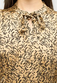 Μπεζ μπλούζα με ακανόνιστο μαύρο γεωμετρικό σχέδιο και δεμένο φιόγκο στο ντεκολτέ σε ένα άτομο με μακριά σκουρόχρωμα μαλλιά.