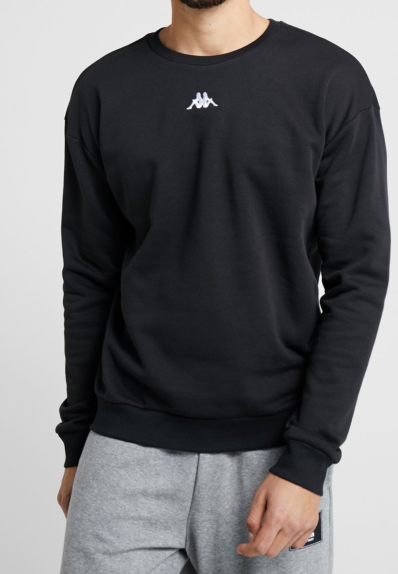 Man som bär en svart långärmad sweatshirt med en liten vit logotyp centrerad nära kragen, matchad med ljusgråa sweatpants.