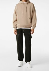 Beige hoodie med ficka fram, tillsammans med svarta cargobyxor som har sidofickor och kontrasterande sömmar, bärs med vita skor.