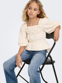 Gaisīgi bēša, teksturēta blūze ar kvadrātveida izgriezumu un pūstām piedurknēm, kopā ar ziliem džinsiem ar baltām plankumiem.