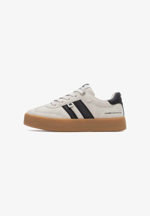 Zapatillas de ante gris claro con rayas negras, puntera redondeada y suela de goma color caramelo. Presenta cordones y detalles de logotipo en el lateral.