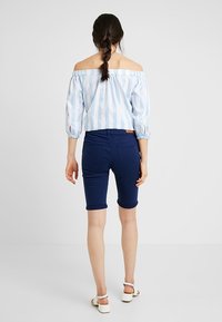 Blusa de rayas azul claro y blanca, sin hombros, con mangas abullonadas, combinada con pantalones cortos hasta la rodilla azul marino. Sandalias blancas de tiras.