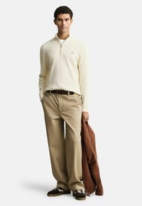 Helles, beiges Strickpullover mit einem halben Reißverschluss, tanfarbene Hose, brauner Gürtel und braune Jacke über einem Arm. Schwarz-weiße Sneaker.