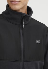 Chaqueta negra con cremallera y cuello de forro polar. Presenta paneles superiores lisos, un parche de logo cosido y una textura de forro polar contrastante en la parte inferior.