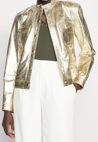 Metalická zlatá bunda s hadím vzorem, s límečkem, předními kapsami a knoflíkovým zapínáním, kombinovaná s tmavě zeleným topem a bílými kalhotami.