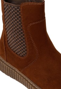 Brown suede ankle boot with textured woven panel, contrasting stitching, and rubber sole featuring a ridged pattern. Side elastic for fit.