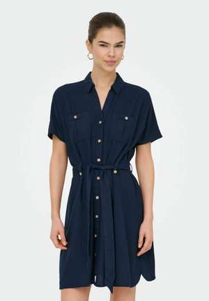 Femme portant une robe chemise bleu marine à manches courtes avec des poches avant et une ceinture à la taille, debout devant un fond uni.