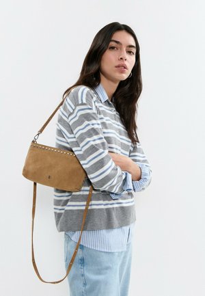 Mujer con cabello largo y oscuro que lleva un suéter gris y blanco a rayas sobre una camisa azul a rayas, vaqueros claros y un bolso de hombro de ante marrón.