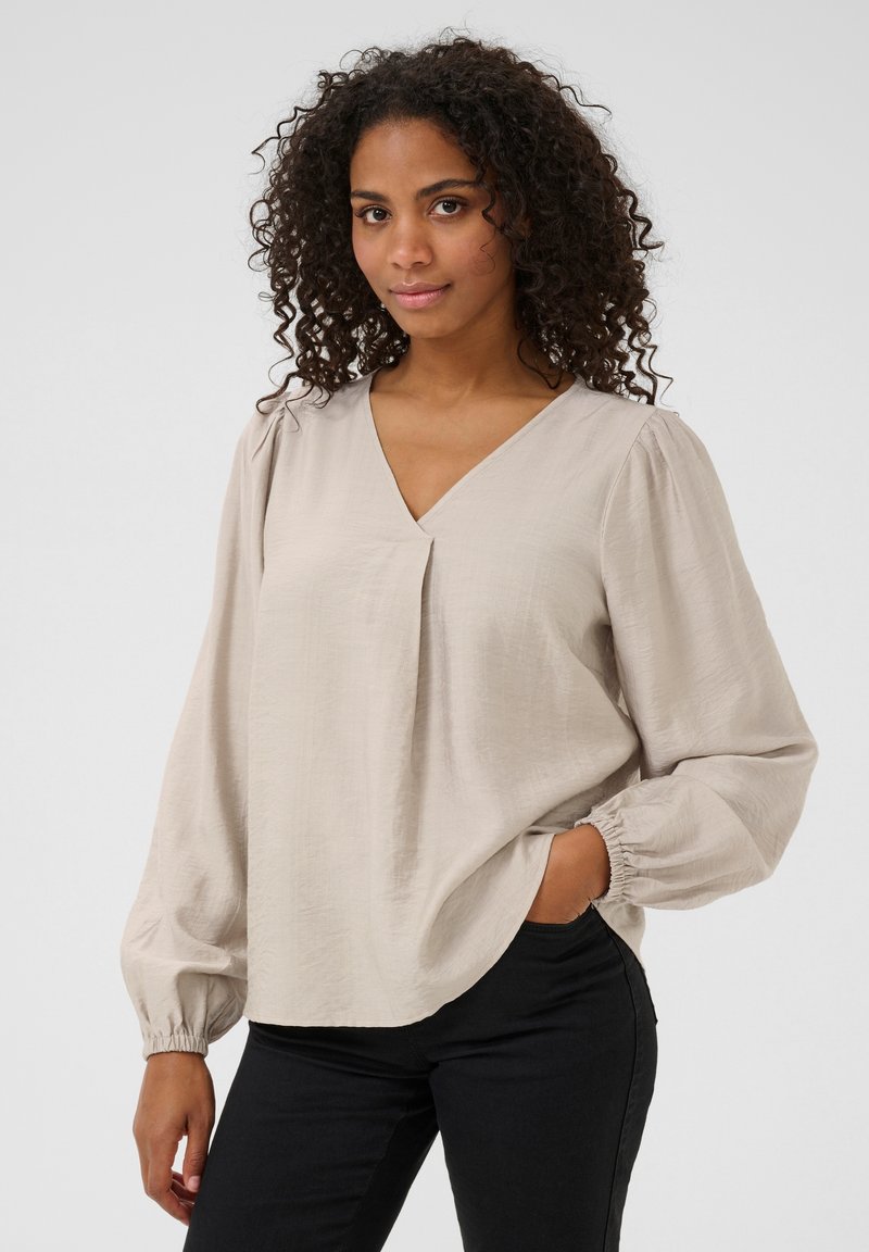 Blouse beige clair avec un décolleté en V, des détails plissés et de longues manches bouffantes avec des poignets élastiques. Associée à un pantalon noir. Texture lisse.