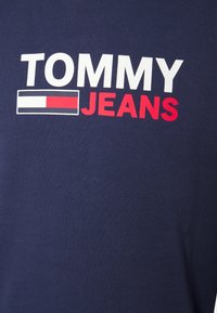 Mörkblått tyg med texten "TOMMY JEANS" i vitt och rött, åtföljd av en rektangulär logotyp med vita, röda och svarta block.