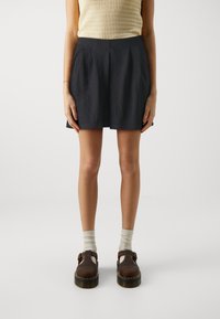 Jupe plissée bleu foncé à taille haute. Portée avec des chaussettes de couleur claire et des sabots marron dotés d'une boucle et d'une semelle épaisse.