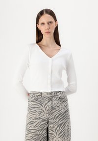 Cardigan blanco acanalado con escote en V y botón frontal; combinado con jeans de estampado de cebra gris. Tejido suave, mangas largas, diseño ajustado.