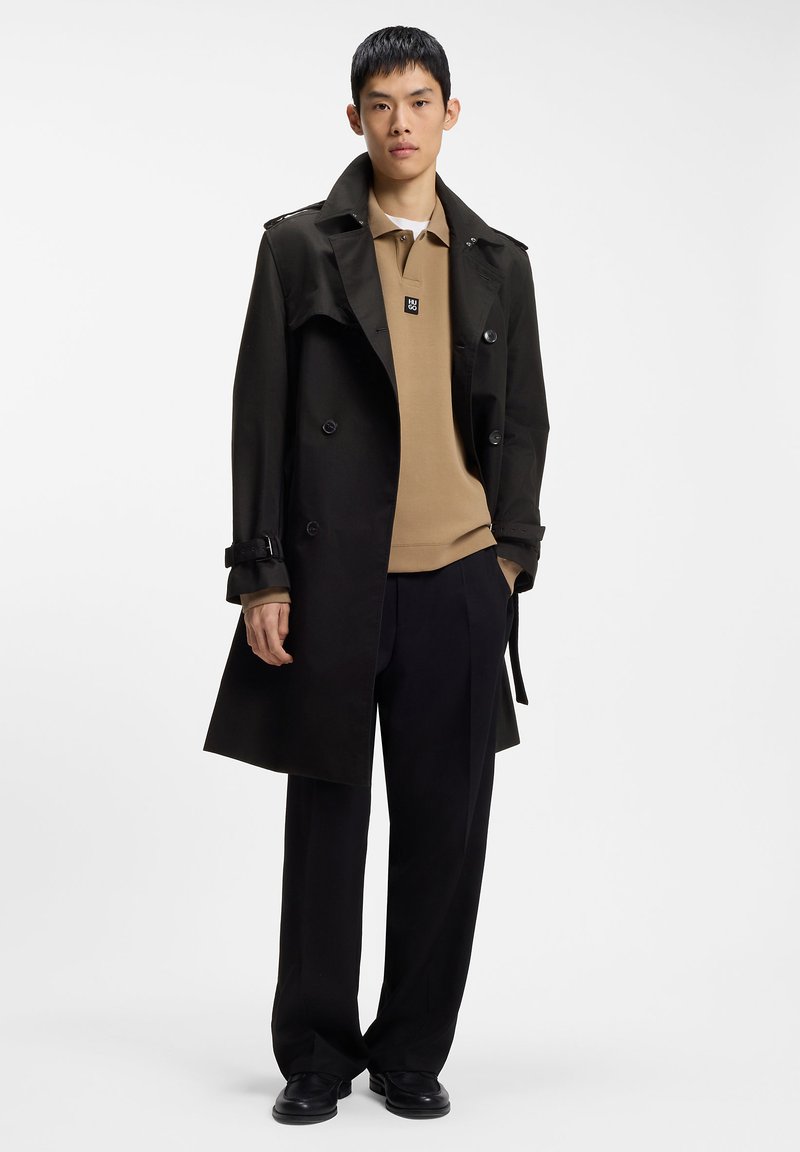 Jeune homme portant un trench-coat noir par-dessus un polo beige, pantalon noir et chaussures noires, debout devant un fond blanc uni.
