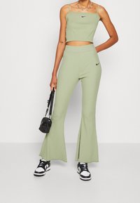 Ljust grönt ribbat crop top med tunna axelband och matchande ribbade utsvängda byxor. Svart Nike-logotyp på toppen och byxorna; modell som håller en liten svart väska.