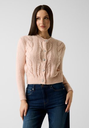 Donna con capelli lunghi e scuri che indossa un cardigan rosa pallido a trecce con bottoni argentati e jeans a vita alta blu scuro, in piedi davanti a uno sfondo semplice.