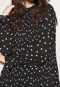 Svart plisserad blus med långa, puffiga ärmar prydda med vita prickar. Rundad halsringning med subtila veck. Mjuk, lättvikts tyg.