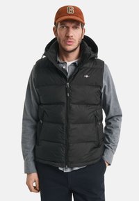 Svart pufferjacka med dragkedja fram, sidofickor och huva. Bärs över en grå skjorta med krage och kompletteras av en orange keps.