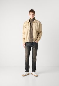 Chaqueta bomber beige, suéter de cuello alto marrón, jeans azules, zapatillas blancas con acentos beige. Tejido ligero, puños elásticos, ajuste casual.