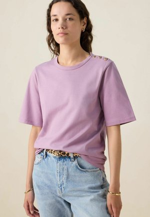 T-shirt en coton lilas à manches courtes, encolure ronde et accents de boutons dorés décoratifs sur l'épaule, associé à un jean bleu clair.
