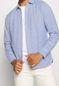 Modrá a bílá pruhovaná košile s knoflíky vyrobená z lehké tkaniny, s klasickým límcem, dlouhými rukávy a manžetami na knoflíky.