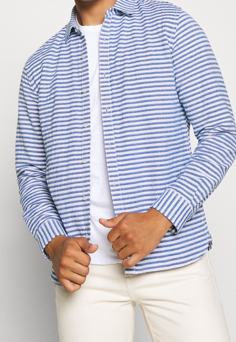 Modrá a bílá pruhovaná košile s knoflíky vyrobená z lehké tkaniny, s klasickým límcem, dlouhými rukávy a manžetami na knoflíky.