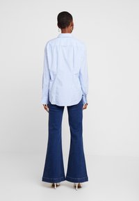 Blauw-wit gestreept overhemd met knopen, lange mouwen, in een relaxte pasvorm, gecombineerd met donkerblauwe uitlopende jeans en witte schoenen met hak.