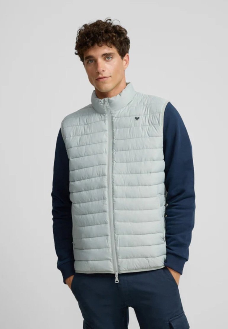 Chaleco acolchado gris claro con un cuello alto, cierre de cremallera y diseño de acolchado horizontal; acompañado de mangas en azul marino. Suave y de textura lisa.